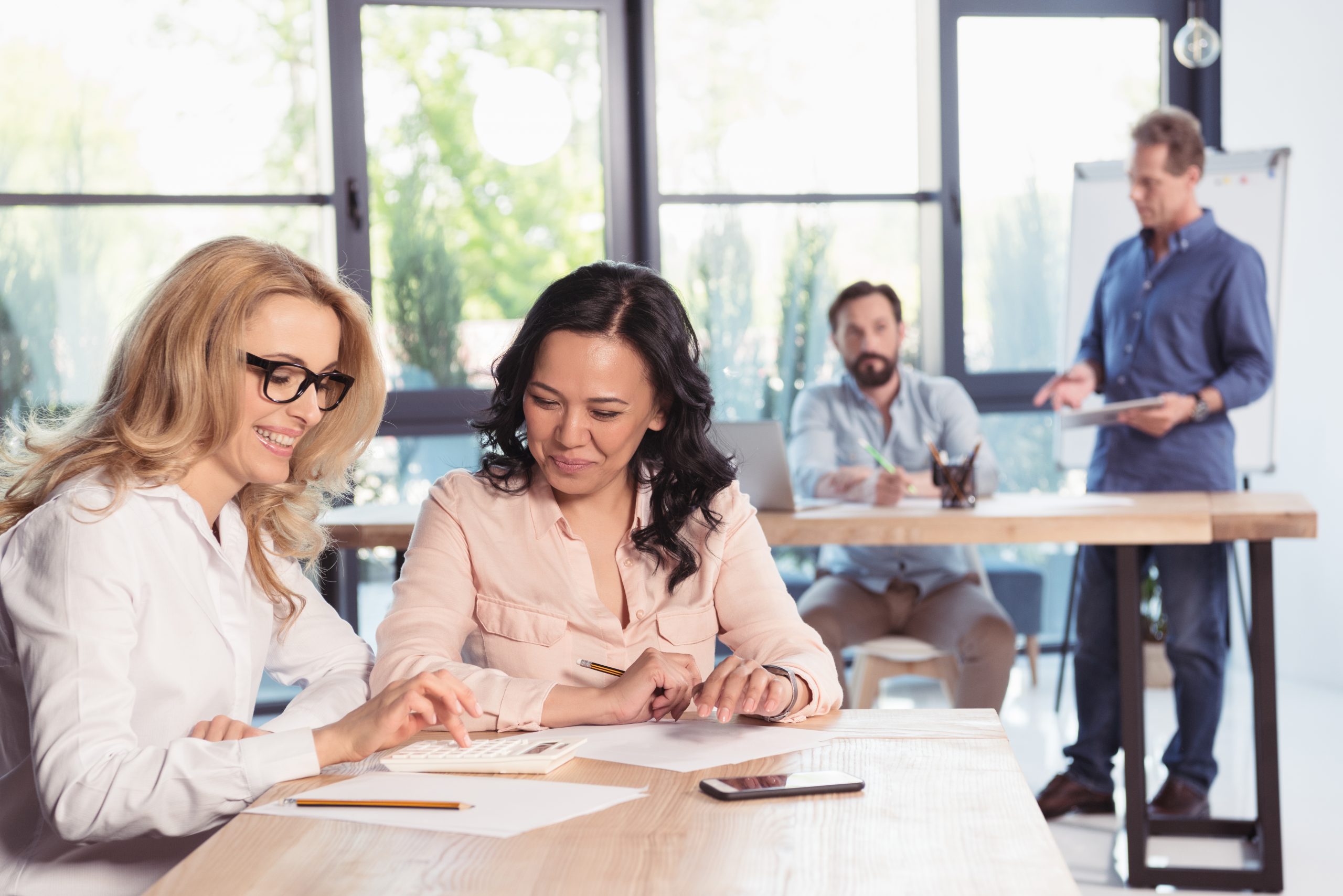 Two employees sit together at a desk and go over employee benefits, while other employees are behind them in the background.