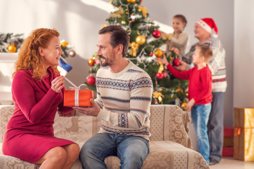 A wife cheerfully opens a Christmas gift presented to her by her husband. The children and grandfather are in the background playing with the Christmas tree.