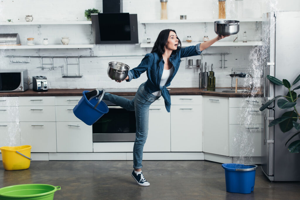 A woman is holding a bowl with her left hand trying to catch water that is falling from her ceiling. She is holding a bowl in her right hand and has a bucket on her foot to help catch the water.