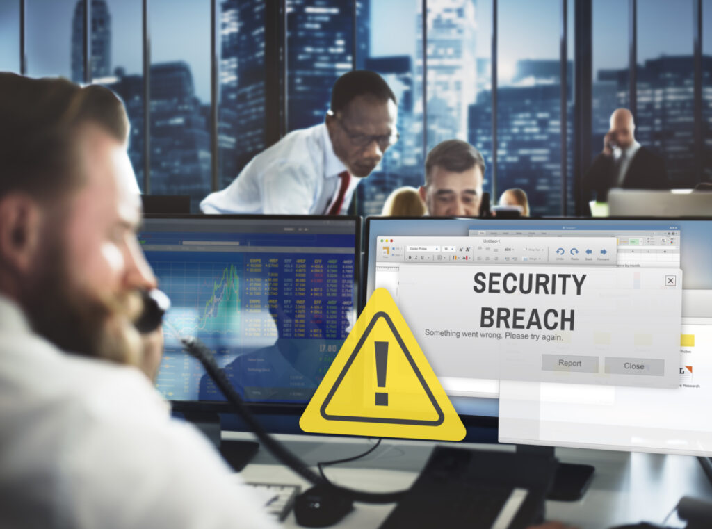 An office worker is on the phone as he's dealing with a security breach to his company's system. The words "SECURITY BREACH" pop up on his computer screen.